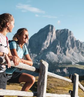 Zwei Wanderinnen lehnen am Holzzaun und genießen den Blick auf markante Berggipfel im Sommer.